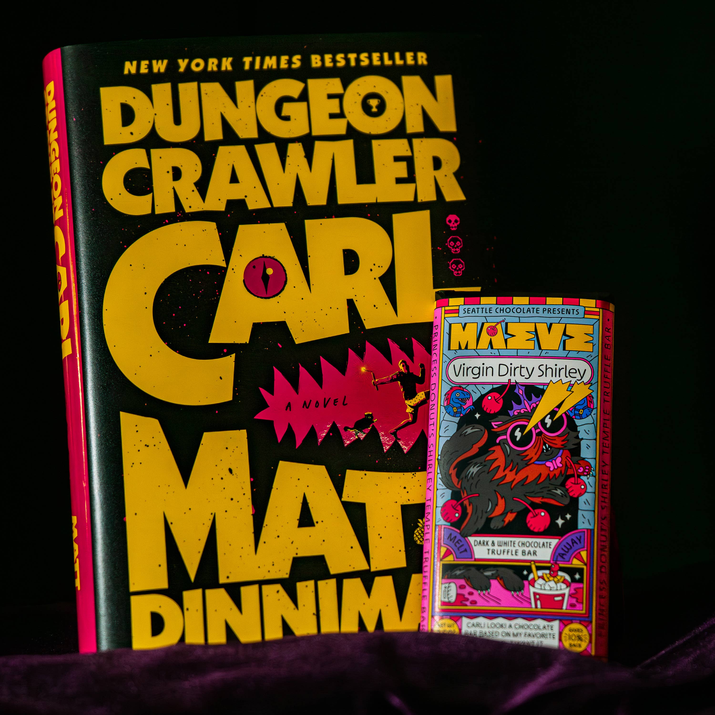 A book titled "Dungeon Crawler" by Carl Mattinelli is displayed upright next to a vintage-style comic book cover on a dark background with a purple fabric surface at the bottom.